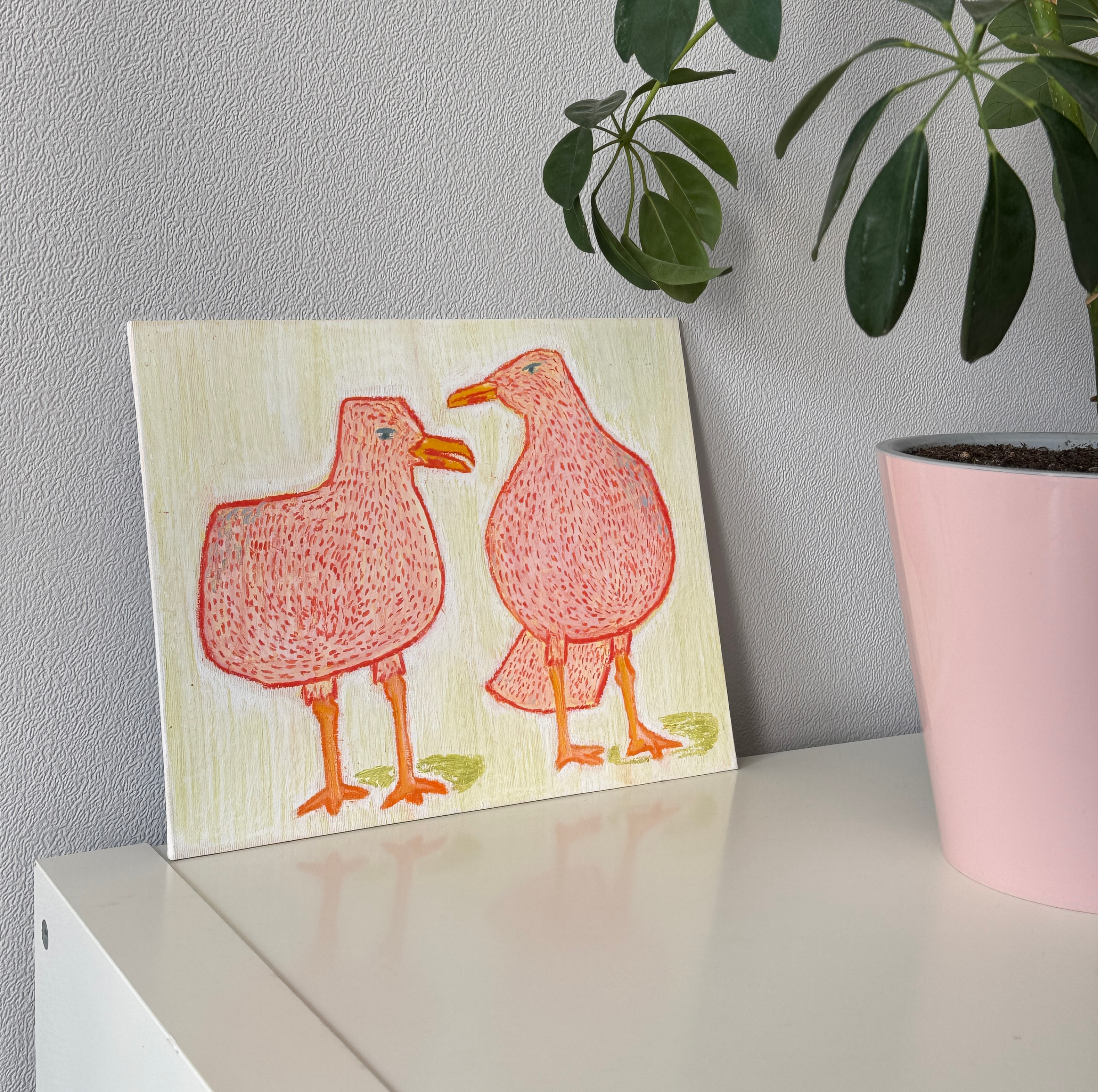 Картина, выполненная масляной пастелью и карандашами на картоне, с изображением двух розовых птиц на светлом фоне стоит на белой полке рядом с розовым горшком с зеленым растением на фоне серой стены