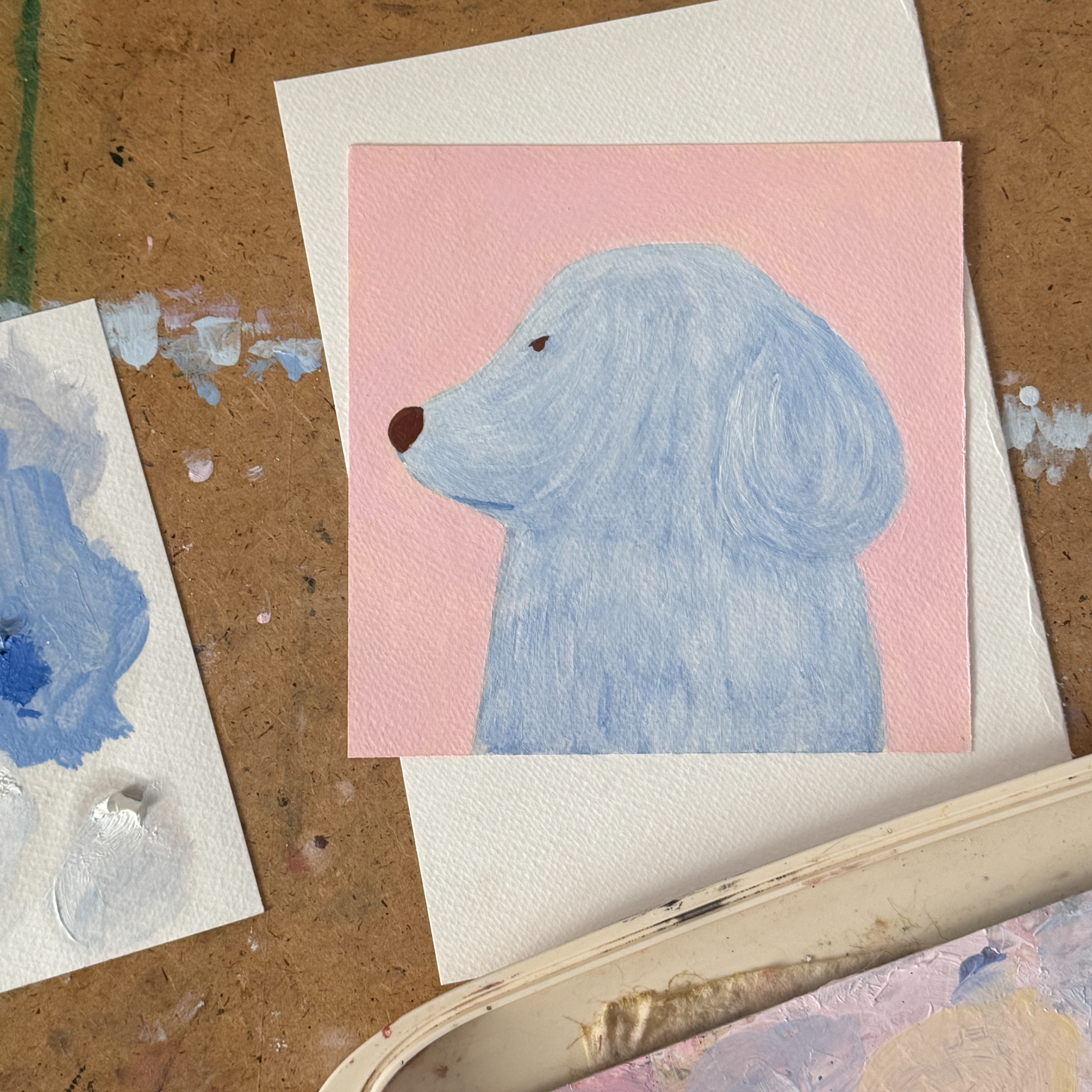 Картина маслом на бумаге с изображением светло-голубой собаки в профиль на розовом фоне лежит на белой бумаге на деревянном столе с пятнами краски, рядом видны палитра и тестовые мазки, бумага
