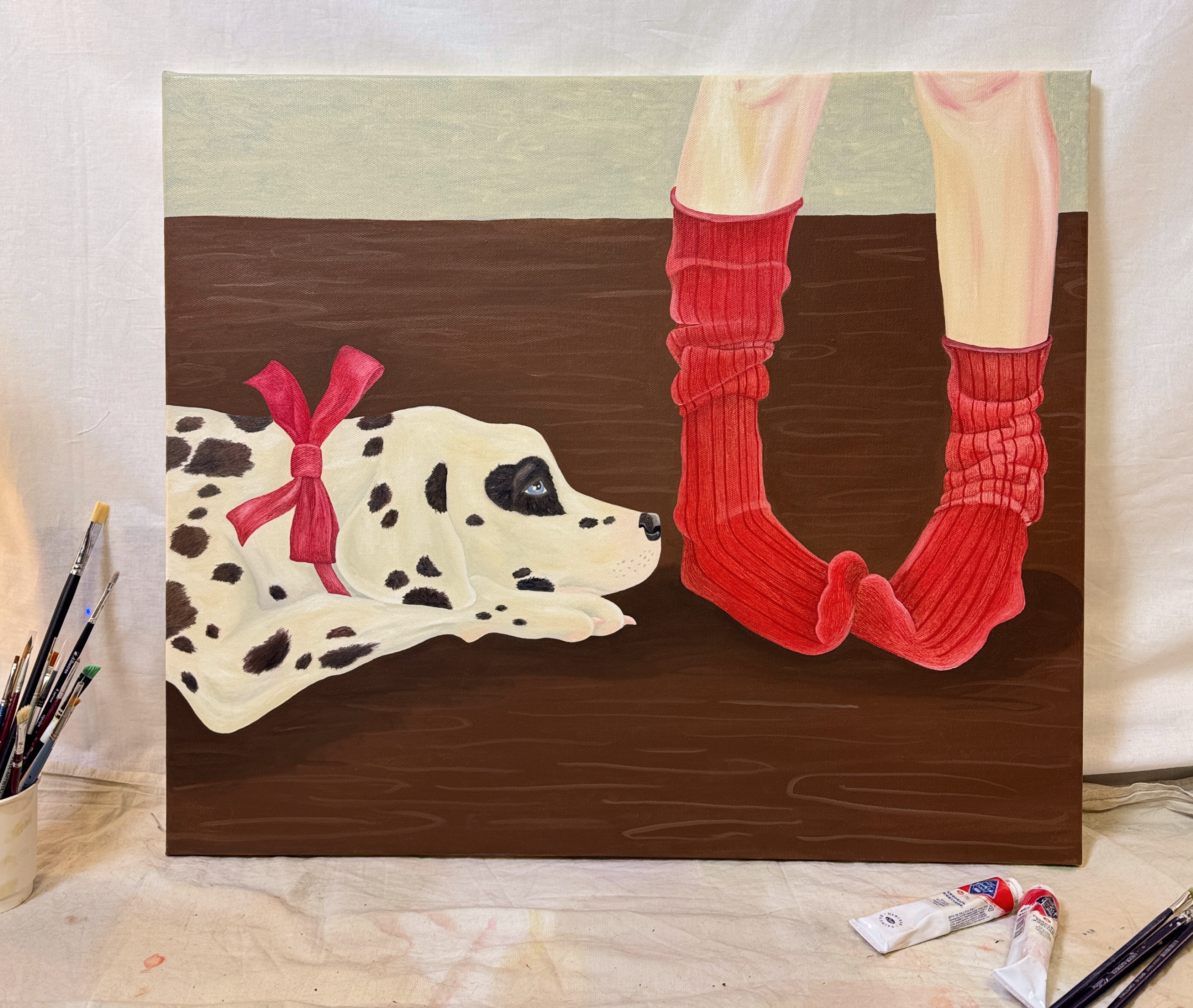 Картина маслом на холсте с лежащим далматином в красном банте и двумя ногами в красных носках лежит на белой ткани среди кистей и тюбиков краски