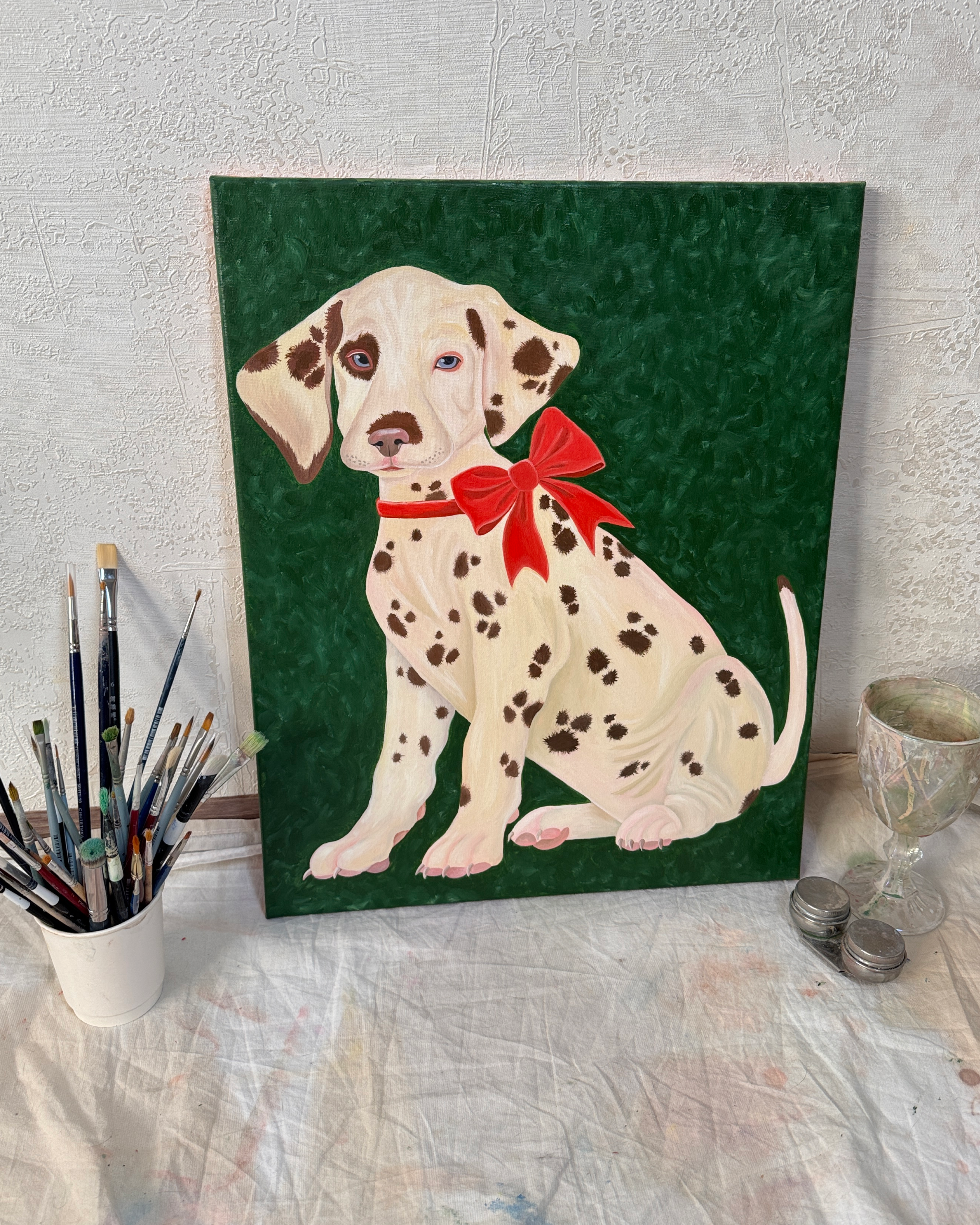 Картина маслом на холсте с сидящим щенком далматинца в красном банте на зеленом фоне стоит у белой стены, окруженная стаканом с кистями, стеклянным бокалом и металлическими баночками на покрытой краской белой ткани
