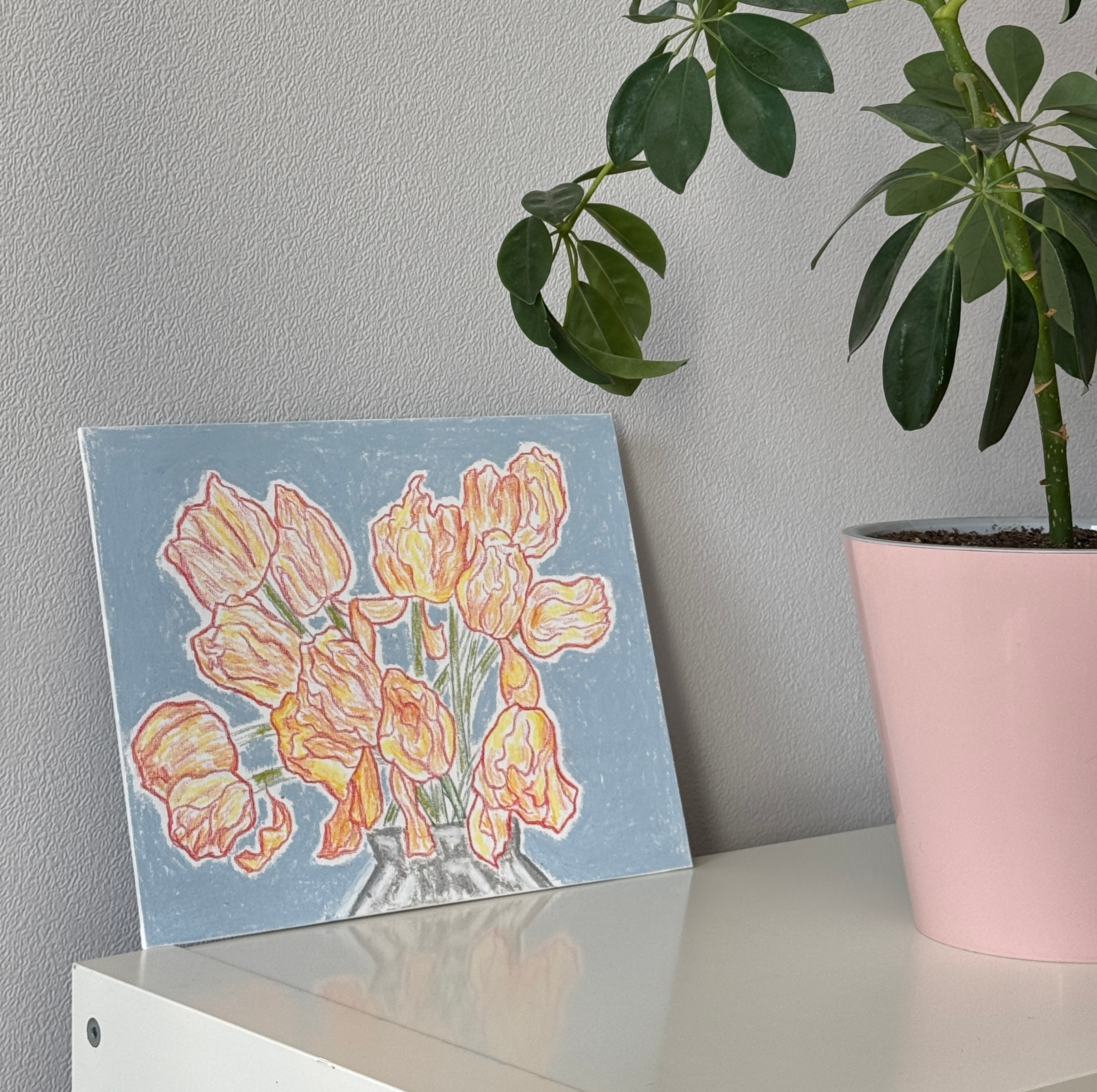 Картина с букетом желтых тюльпанов в серой вазе на голубом фоне, выполненная масляной пастелью и карандашами на картоне, стоит на белой полке рядом с зеленым растением в розовом горшке на фоне серой стены